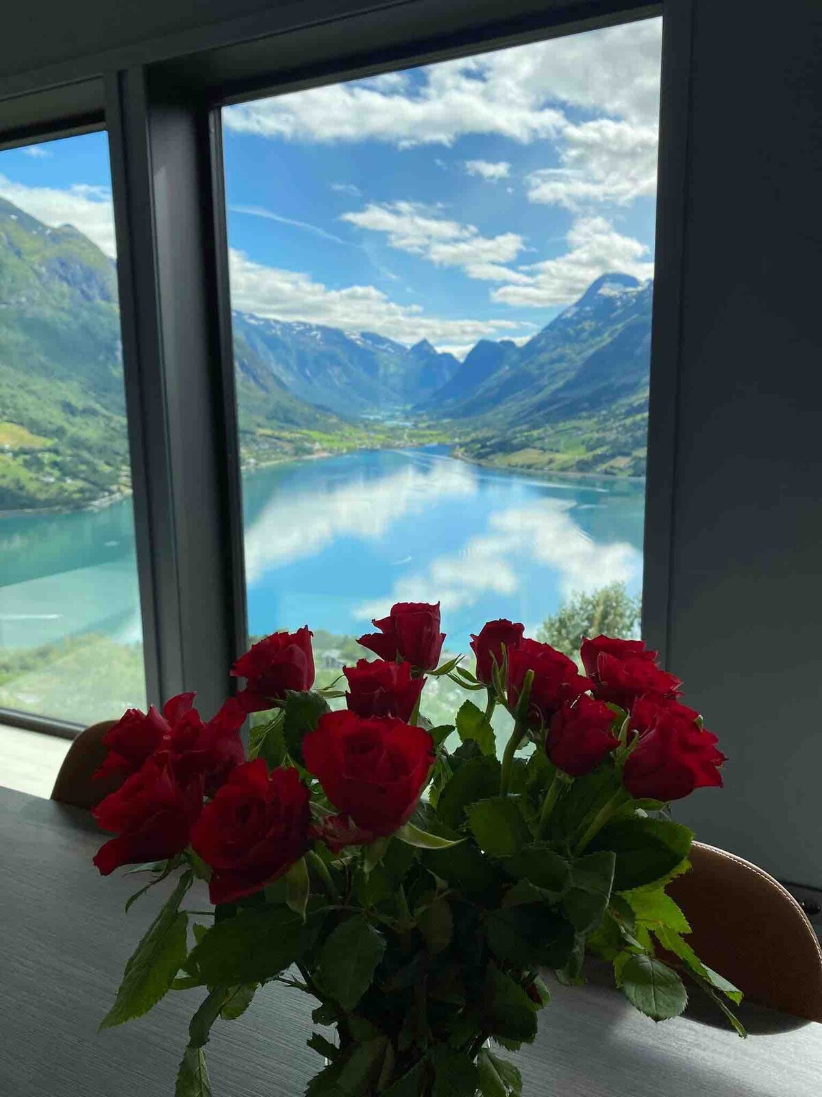 A vase of red roses is positioned on a table, with a stunning view of the fjord and mountains visible through large glass windows. The landscape reflects in the water, creating a serene backdrop, while clouds drift above the peaks.