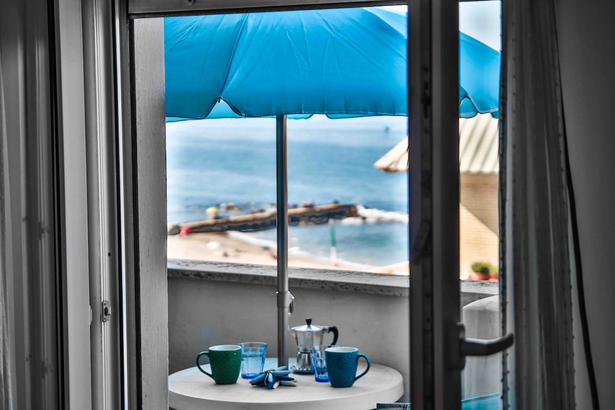 A small balcony with a round table is visible, featuring two green cups and a glass. An espresso maker rests on the table, complemented by a blue umbrella providing shade. The serene sea and distant coastline can be seen through the open balcony door.