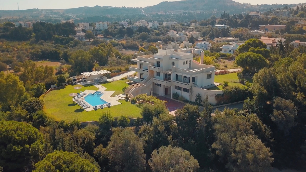 Aerial view of the villa showcases a spacious outdoor area with a private swimming pool and lounge chairs. Lush green lawns surround the property, complemented by well-kept gardens. The villa’s modern architecture is highlighted under a clear sky, emphasizing its tranquil and secluded location.