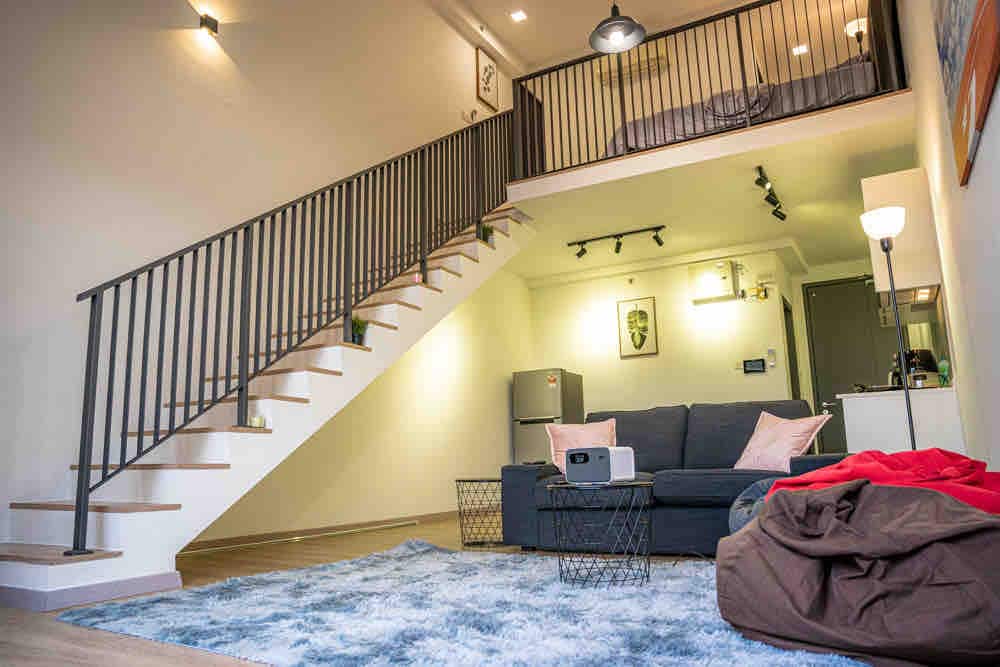 A modern, multi-level living area features a cozy sofa with soft cushions, accompanied by a bean bag chair. A plush area rug adds comfort to the floor. Stairs lead to an upper level where a spacious sleeping area is visible, enhancing the layout of the space.