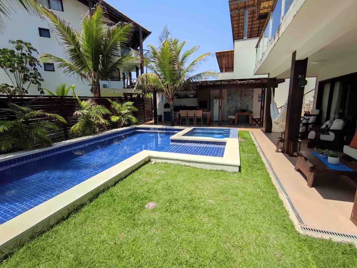 An outdoor area features a rectangular swimming pool surrounded by lush green grass and tropical plants. A smaller jacuzzi is adjacent to the pool. An open-air seating area with a dining table and chairs is visible in the background, along with shaded lounge furniture.