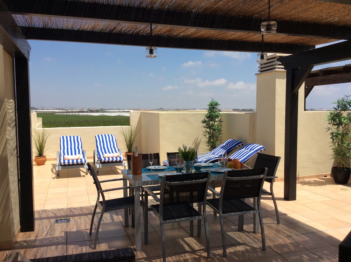 A spacious rooftop terrace features a dining table set for six, surrounded by comfortable lounge chairs with blue and white stripes. Potted plants add greenery, while expansive views of the surrounding landscape are visible in the distance, inviting relaxation and outdoor dining.