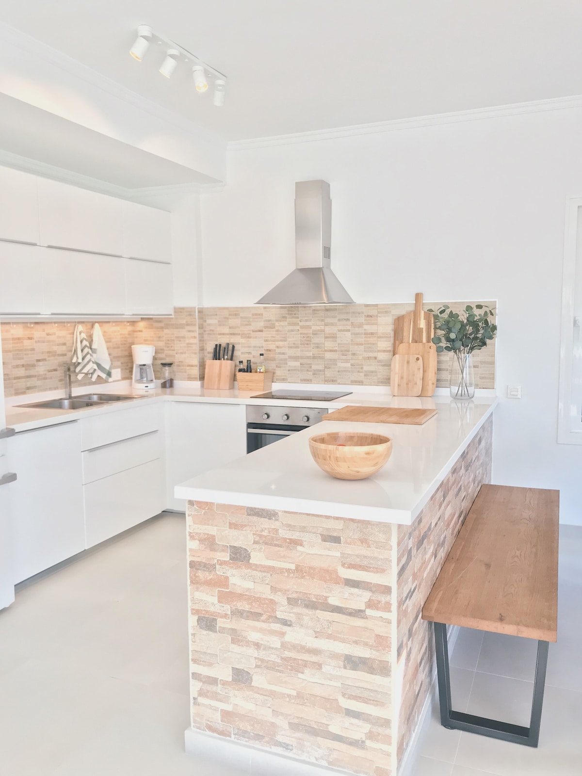 A modern kitchen features sleek white cabinetry and a light-colored stone backsplash. A wooden countertop runs along the kitchen island, which is complemented by a wooden bench. Stainless steel appliances are visible, along with kitchen utensils and decor positioned on the countertop.