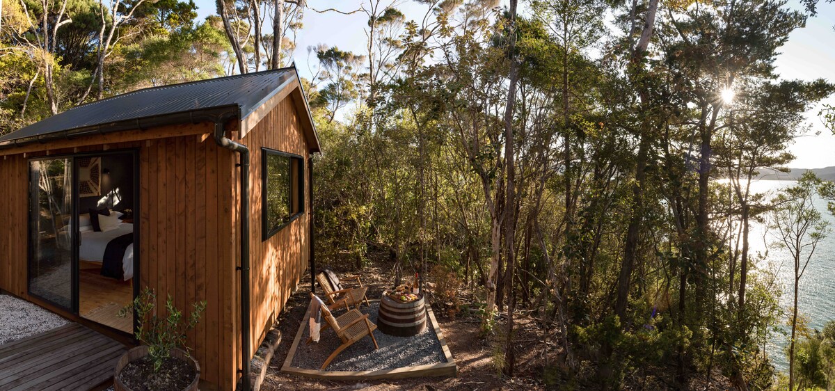 A wooden glamping hut is surrounded by lush native bush, with large glass windows revealing a cozy interior. An outdoor seating area is visible beside a circular bath, offering a serene view of the water through the trees.