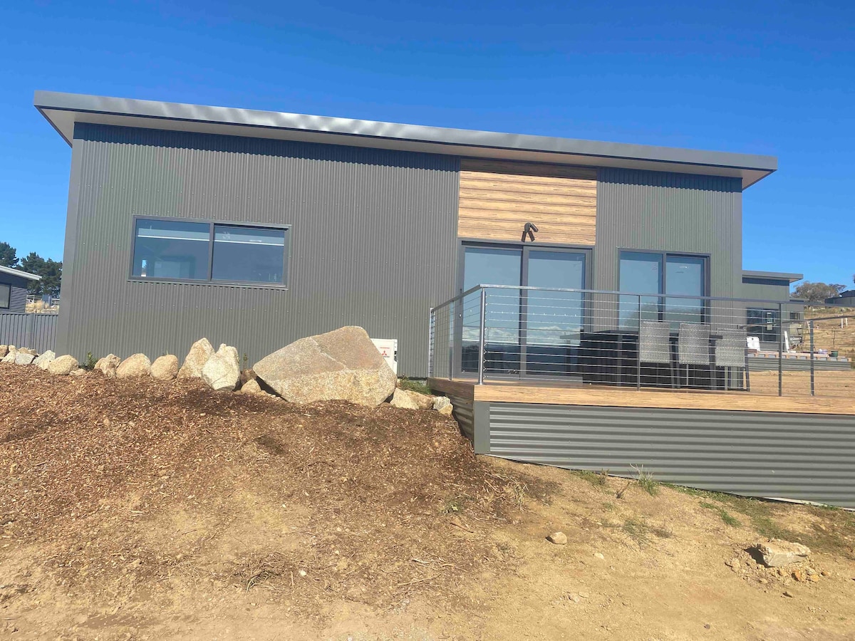 A contemporary cabin exterior is displayed, featuring a combination of corrugated metal and wooden accents. Expansive windows provide views of the surroundings, while a wooden deck with a railing extends from the front. A natural landscape of rocks and soil completes the setting.