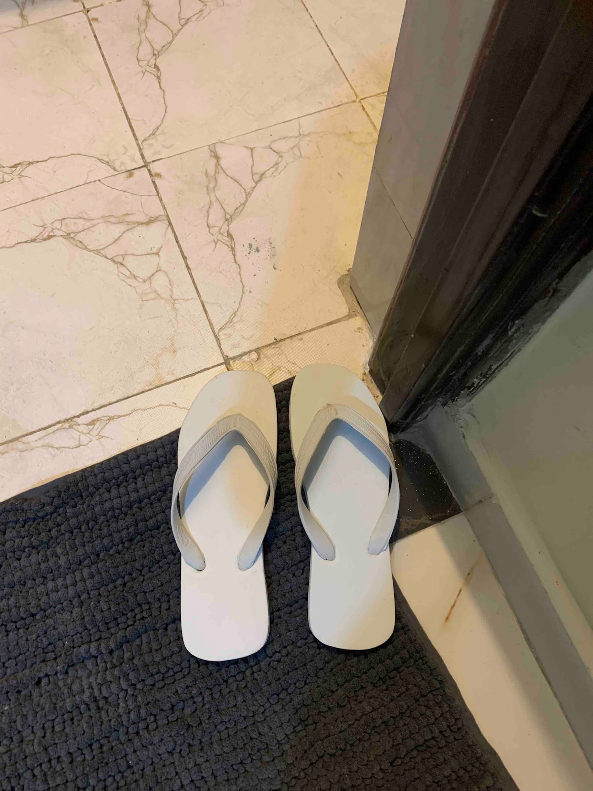 A pair of white flip-flops is placed neatly on a dark blue mat at the entrance of a bathroom. The floor features light-colored, marbled tiles that contrast with the mat's texture.