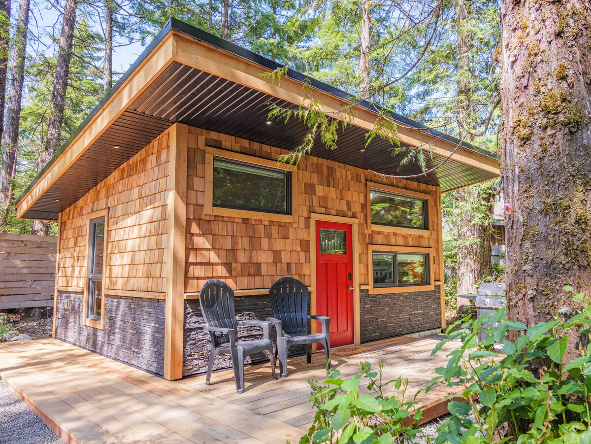 A cedar cabin is surrounded by trees, showcasing a distinct design with wood shingles and stone accents. Large windows provide natural light, while a bold red door adds a welcoming element. Two black chairs are positioned on the wooden deck, inviting relaxation.