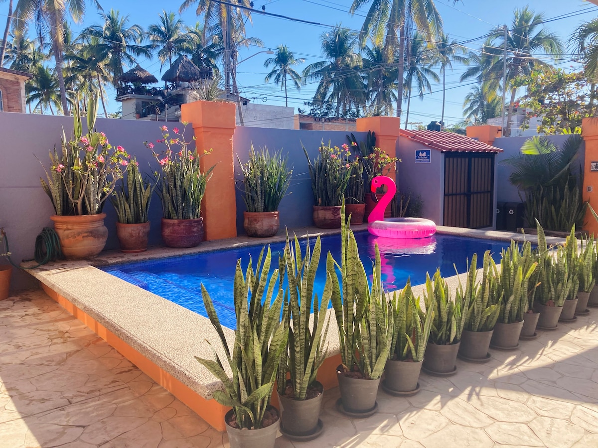 A private pool is surrounded by vibrant potted plants and palm trees, creating a relaxing outdoor space. A large pink float, shaped like a flamingo, adds a playful touch to the water's blue hue. The pool area features a textured stone surface underfoot.