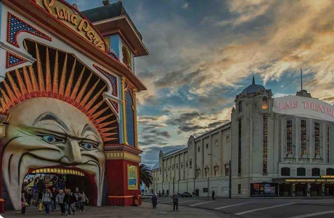 A vibrant entrance to Luna Park features a large, colorful façade with a prominent face design. To the right, a historic theater building showcases classic architecture, illuminated under a dramatic sky filled with clouds. Pedestrians are seen walking, adding life to the bustling scene.