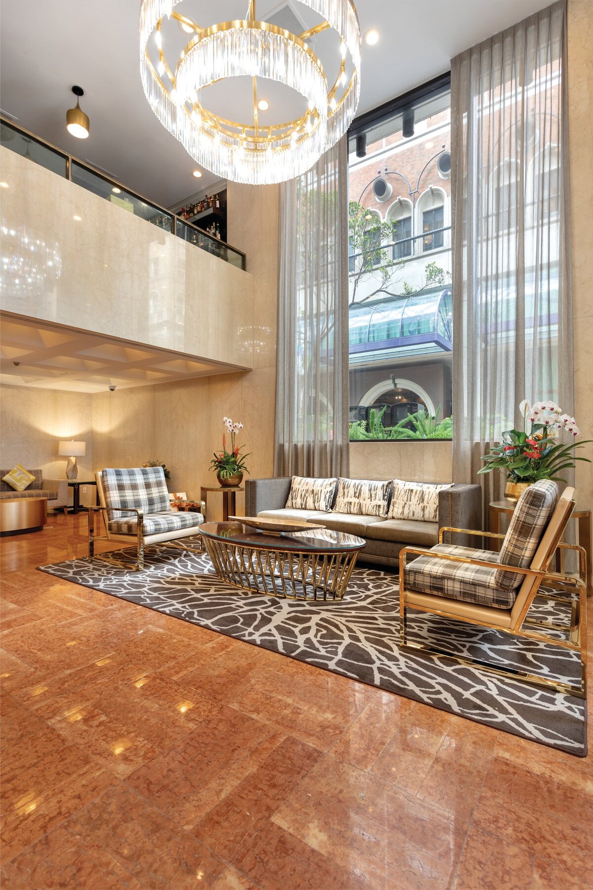 A modern lobby is showcased with large windows allowing natural light to illuminate the space. Comfortable seating is arranged around a central table, complemented by decorative plants. A patterned rug anchors the area, while a stylish chandelier adds an elegant touch above.