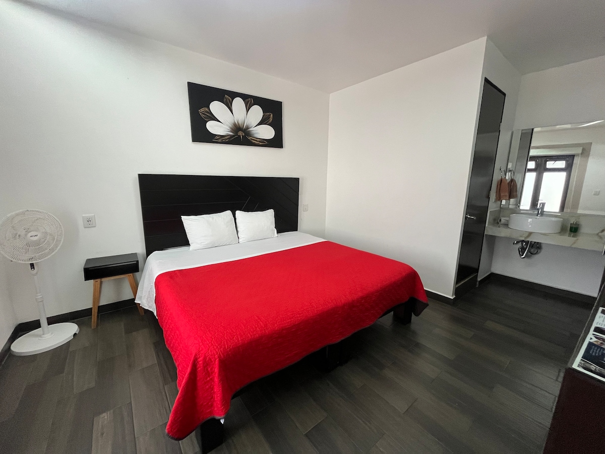 A spacious sleeping area is highlighted by a large red bedspread covering a substantial bed. A sleek black headboard adds a modern touch. A decorative wall art piece featuring a white flower hangs above the bed. The room includes a small bedside table and a fan, with an ensuite bathroom visible in the background.