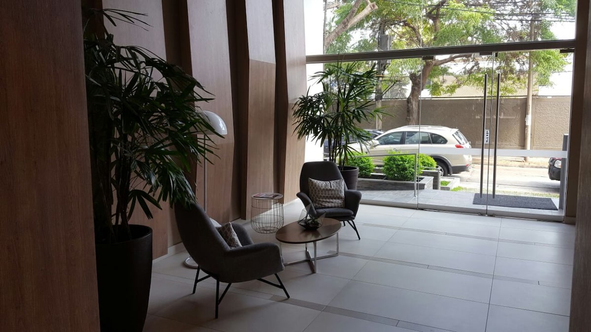 A modern lobby area is depicted, featuring two gray armchairs and a minimalist coffee table. Potted plants add greenery to the space, while large glass doors provide natural light and visibility to the street outside.