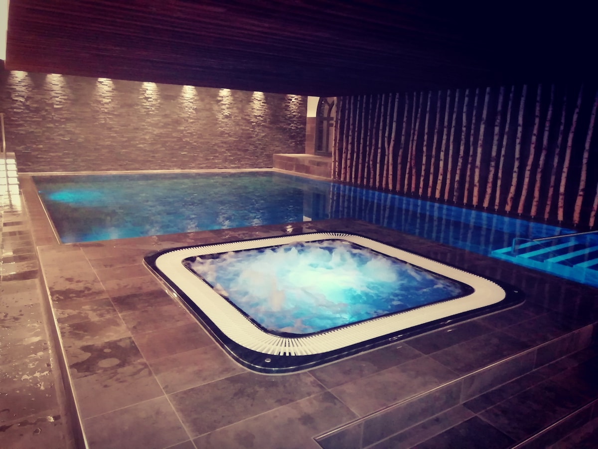 An inviting wellness area features a spacious indoor swimming pool alongside a jacuzzi. The pool's surface reflects soft lighting, while natural stone accents provide a calming backdrop. The contrasting textures of the wood and stone create a serene atmosphere for relaxation and enjoyment.