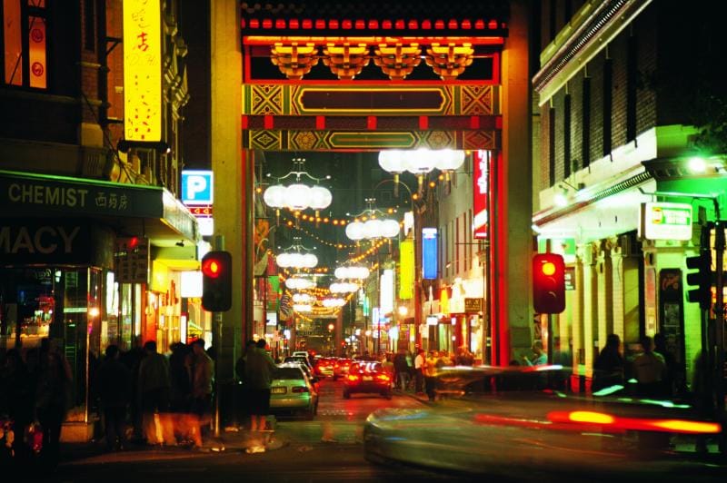 A bustling street scene in Chinatown is captured at night, highlighting bright neon signs and decorative lanterns. Traffic flows along the road, while pedestrians gather under the welcoming glow of storefronts. The iconic archway stands prominently, symbolizing the cultural richness of the area.