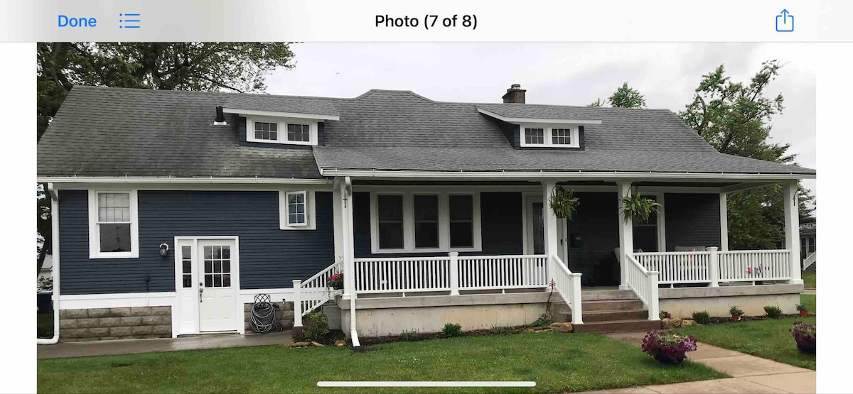 A charming cottage showcases a blue exterior with white trim, featuring a wide front porch adorned with hanging plants. Windows with multiple panes are positioned above and beside the entry door, providing ample natural light. A grassy area is visible in front.