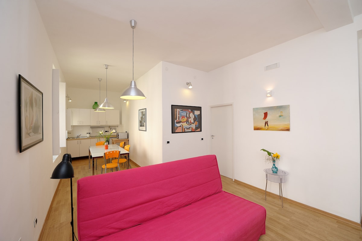 A spacious living area features a bright pink sofa bed and a dining area in the background. The room is illuminated by pendant lights, with modern artwork adorning the walls. A floral arrangement sits on a side table, adding a touch of color.