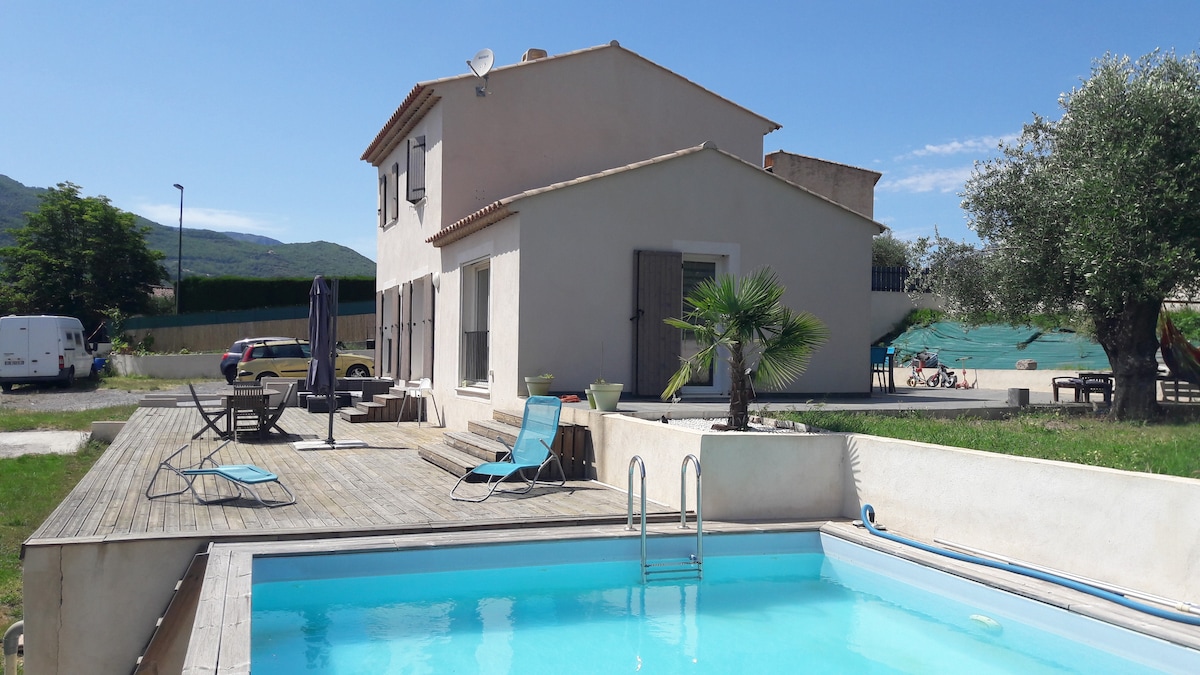 The exterior of the villa is visible, featuring a private pool surrounded by a wooden deck. Sun loungers are placed nearby, and the house showcases large windows under a blue sky. Lush greenery and a distant view of leisure facilities are present in the background.