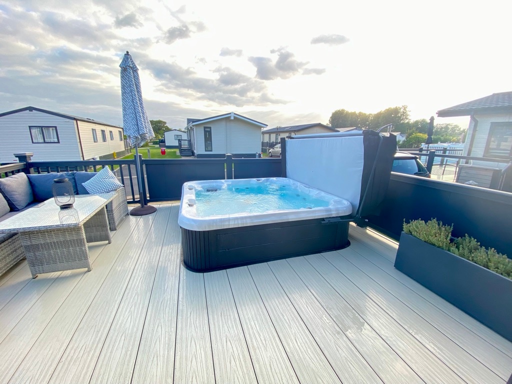 An outdoor decking area features a private hot tub surrounded by a low barrier. A corner sofa with soft cushions provides seating, and a tall umbrella offers shade. Surrounding homes are visible in the background, set against a cloudy sky.