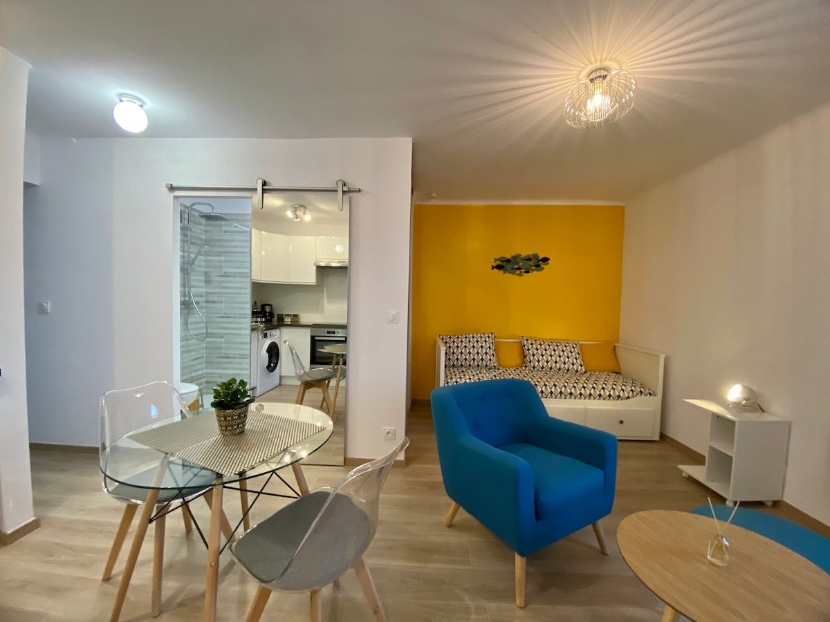 The space features a bright yellow accent wall that enhances the welcoming feel. A glass dining table is surrounded by stylish chairs, while a cozy seating area with a sofa bed and vibrant blue armchairs invites relaxation. Natural light filters through a nearby window.