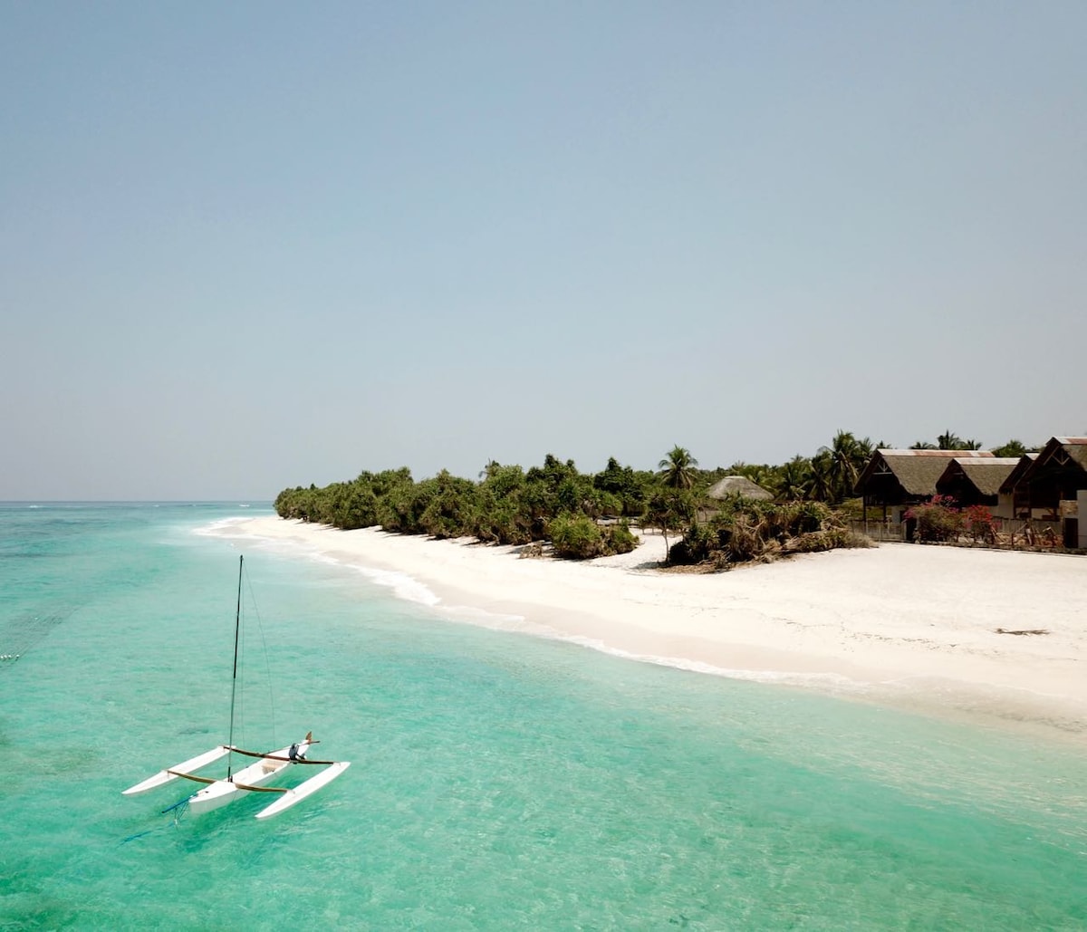 A calm beach scene is depicted, featuring clear turquoise waters gently lapping at the soft white sand. A traditional boat rests on the water, while lush greenery and thatched-roof structures line the shoreline, inviting visitors to enjoy the serene coastal environment.