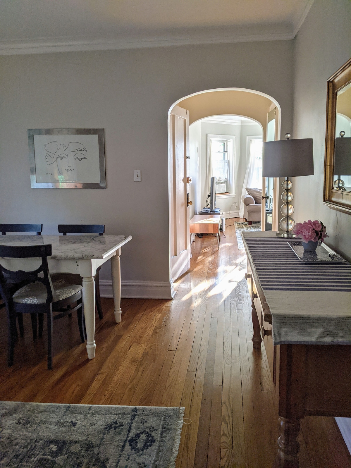 A bright hallway leads from the dining area, featuring a marble-top table surrounded by four black chairs. Natural light filters through a nearby archway, illuminating the wooden floor and inviting atmosphere. A small console table with a lamp and floral decor is positioned against the wall.