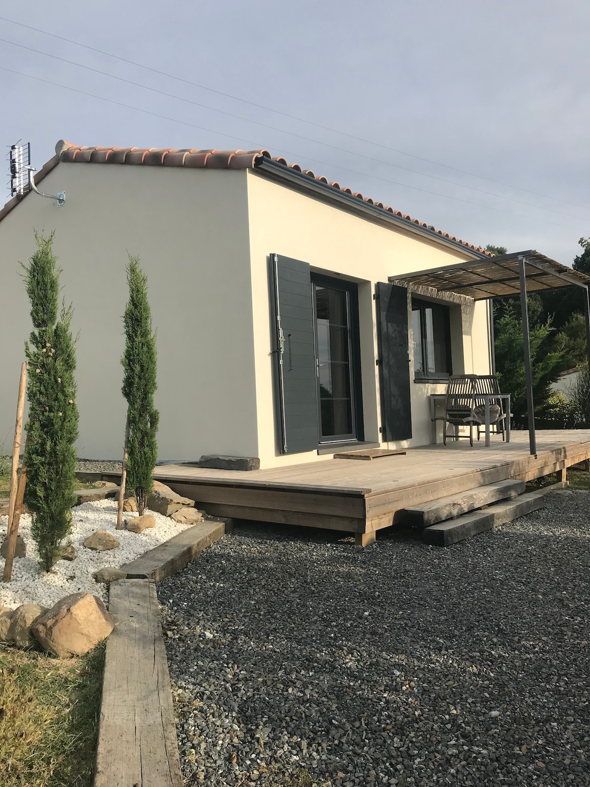 An independent single-story cottage is presented with a wooden deck. Dark shutters adorn the windows, while a small table and chairs invite outdoor seating. Neatly arranged decorative stones and slender plants lead up to the entrance, enhancing the cottage's welcoming appearance.