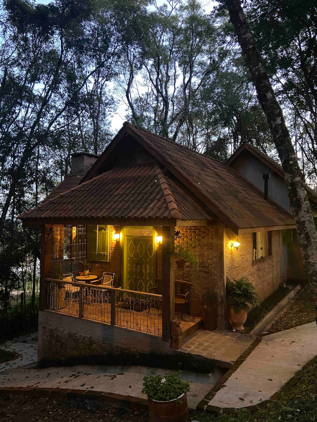 The chalet is nestled among trees, showcasing a warm glow from lanterns illuminating the porch. A welcoming outdoor seating area is visible, with a stone pathway leading to the entrance. The combination of natural surroundings and soft lighting creates an inviting ambiance.