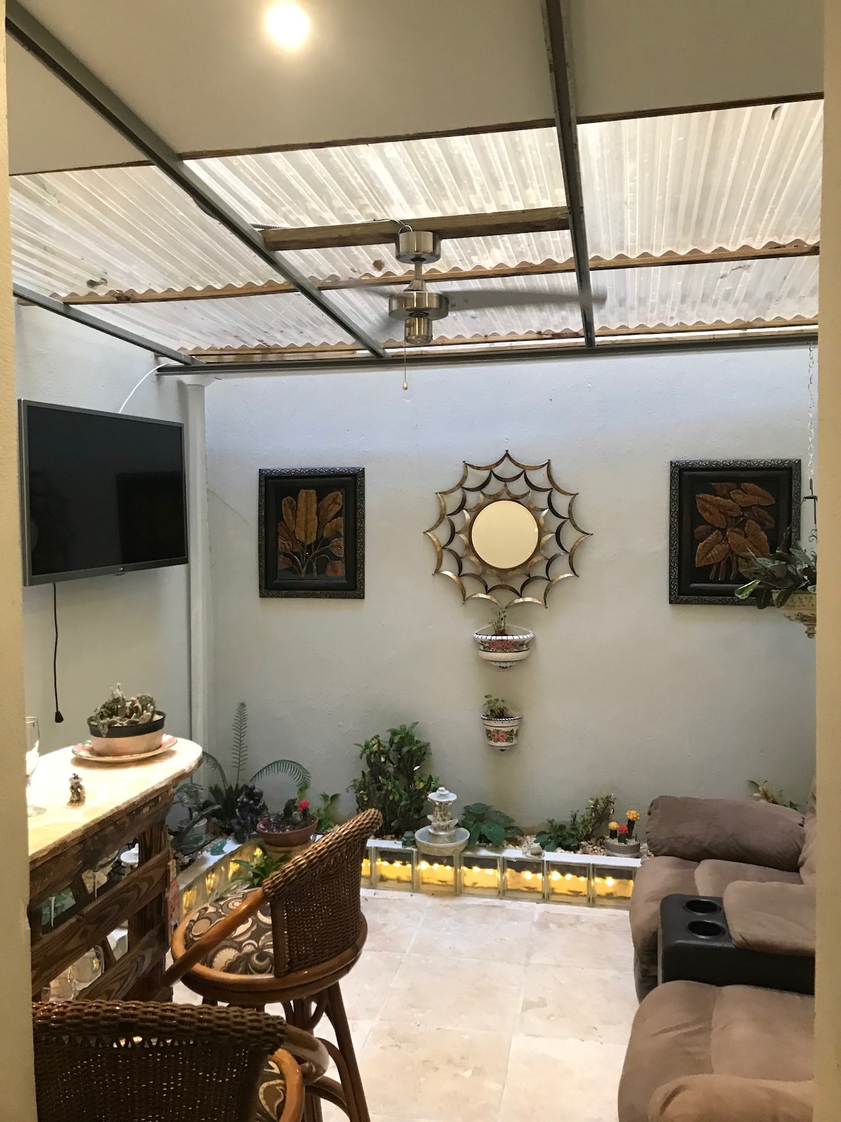 A cozy indoor garden area features a bar with seating and two plush recliners. A small wall-mounted TV is visible. Natural light filters through a clear covered roof, illuminating plants and decor against the muted walls, while a mirror and framed artwork add decorative touches.