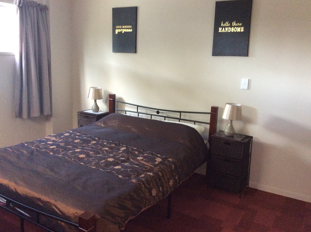A well-appointed bedroom features a queen bed covered in a dark, patterned duvet. Two bedside tables with lamps are positioned on either side. Decorative wall art above the bed adds a touch of character, and light filters in through the window, covered by gray curtains.