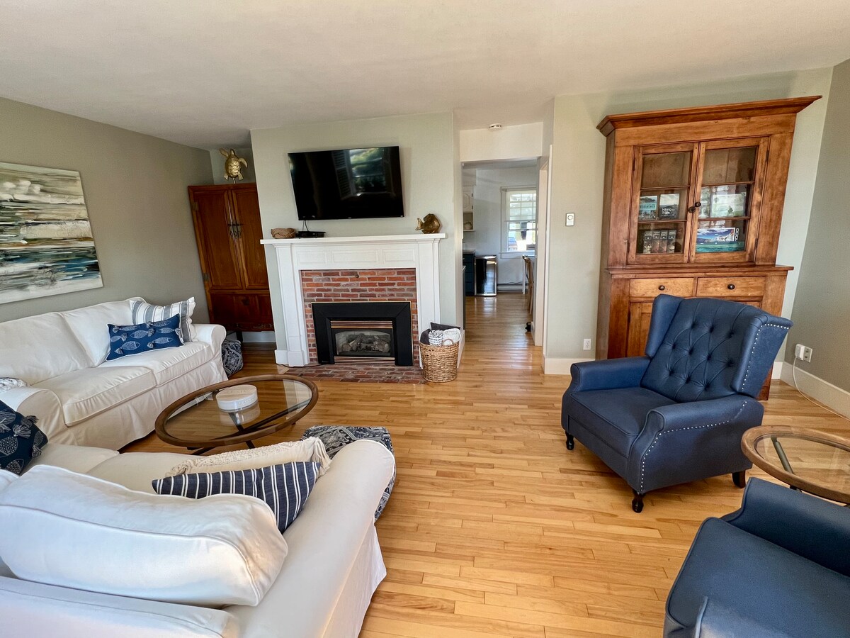 A light-filled living area features two white sofas and a blue accent chair arranged around a glass coffee table. A neutral-toned rug is placed on hardwood flooring. A brick fireplace with a television above adds to the inviting atmosphere, complemented by wooden cabinetry in the background.