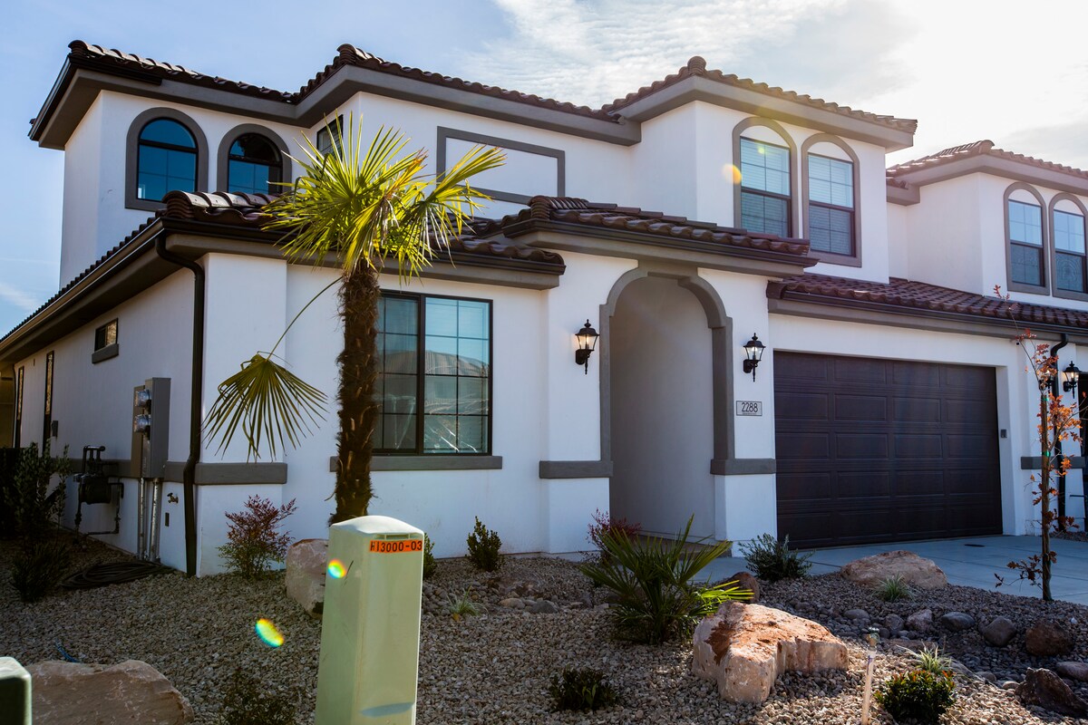 The exterior of a modern home features a stucco façade with a combination of arched and rectangular windows, accented by decorative light fixtures. Lush landscaping with various plants and rocks surrounds the entryway, while a double garage door is visible on the right side.