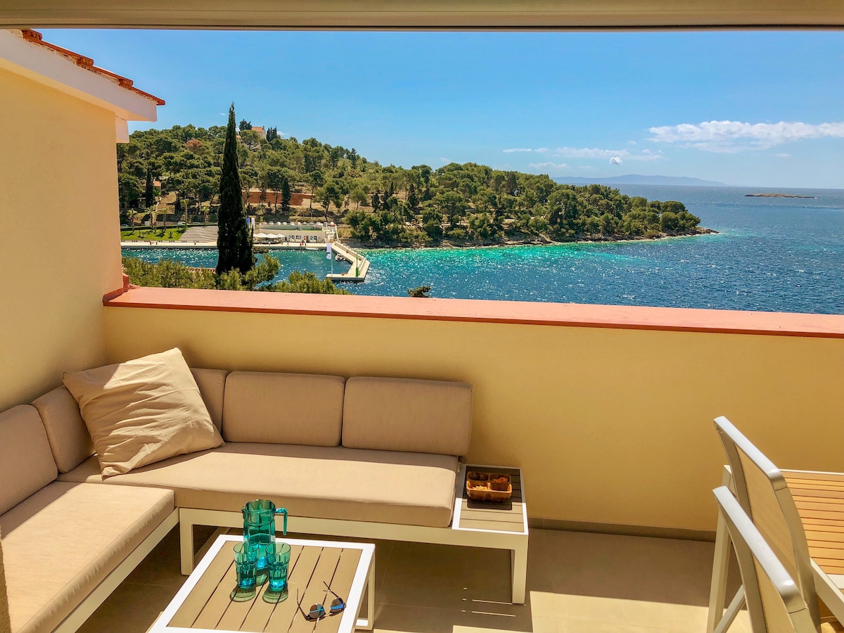 A comfortable balcony area features a spacious outdoor seating arrangement with neutral cushions. A glass coffee table holds decorative items. The backdrop reveals a panoramic view of the sparkling blue sea and lush green coastline, complemented by clear skies.