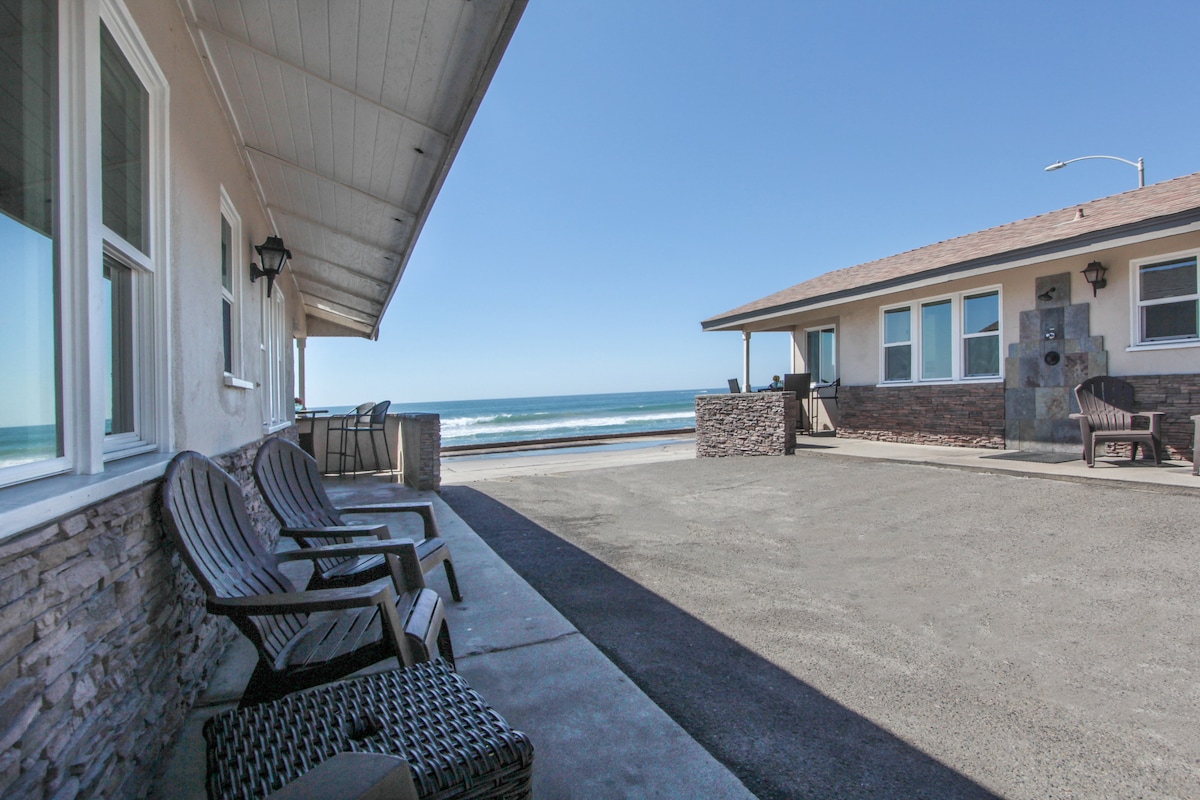 A spacious outdoor area is shown, featuring several wooden chairs positioned along a paved pathway. The ocean is visible in the background, with gentle waves lapping at the shore under a clear blue sky. A neighboring building is present on the right.