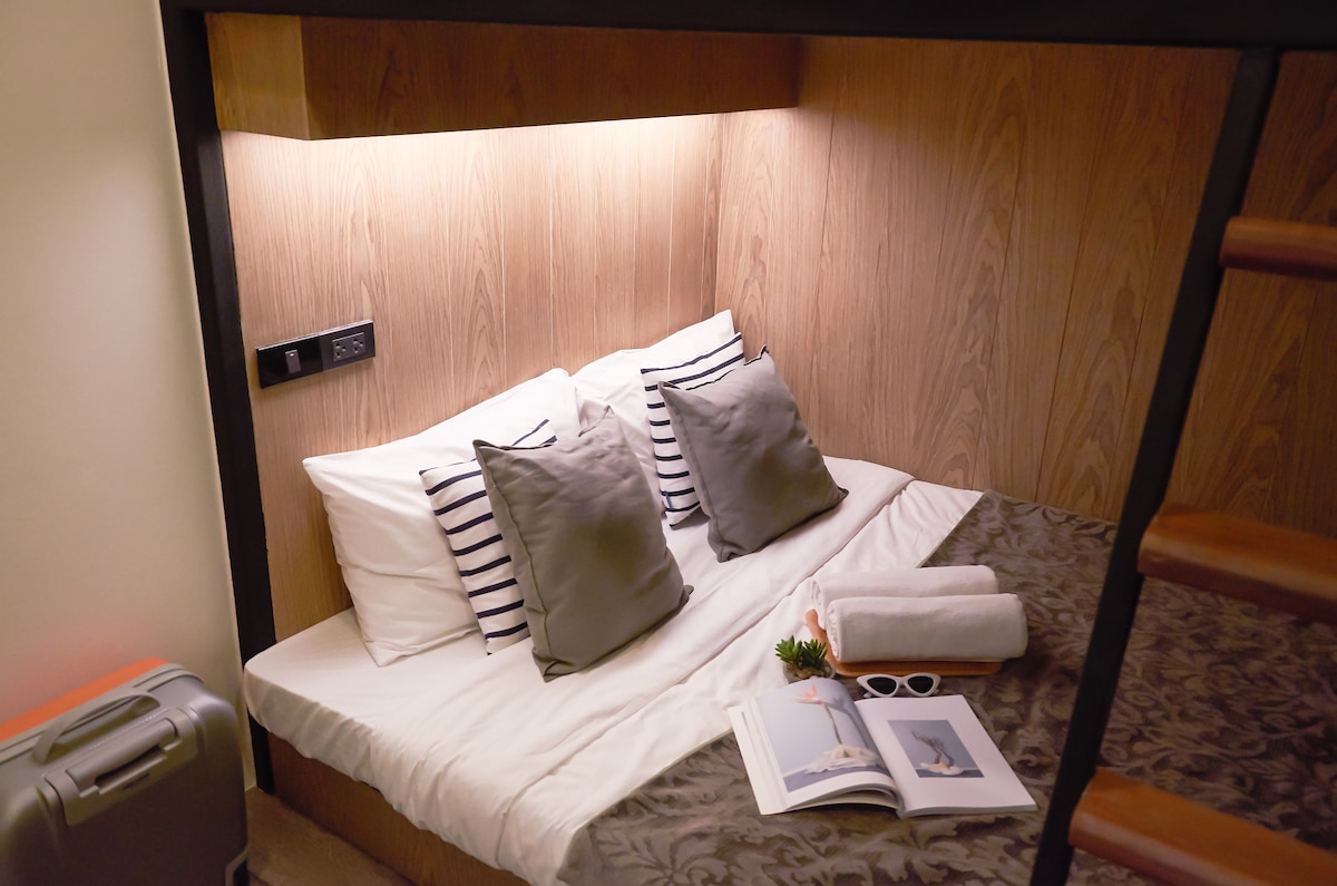 A cozy sleeping area features a plush, spacious bed adorned with striped and solid pillows. Soft lighting highlights the wooden wall paneling, creating a gentle ambiance. A small table is positioned beside the bed, complemented by a suitcase and neatly rolled towels.