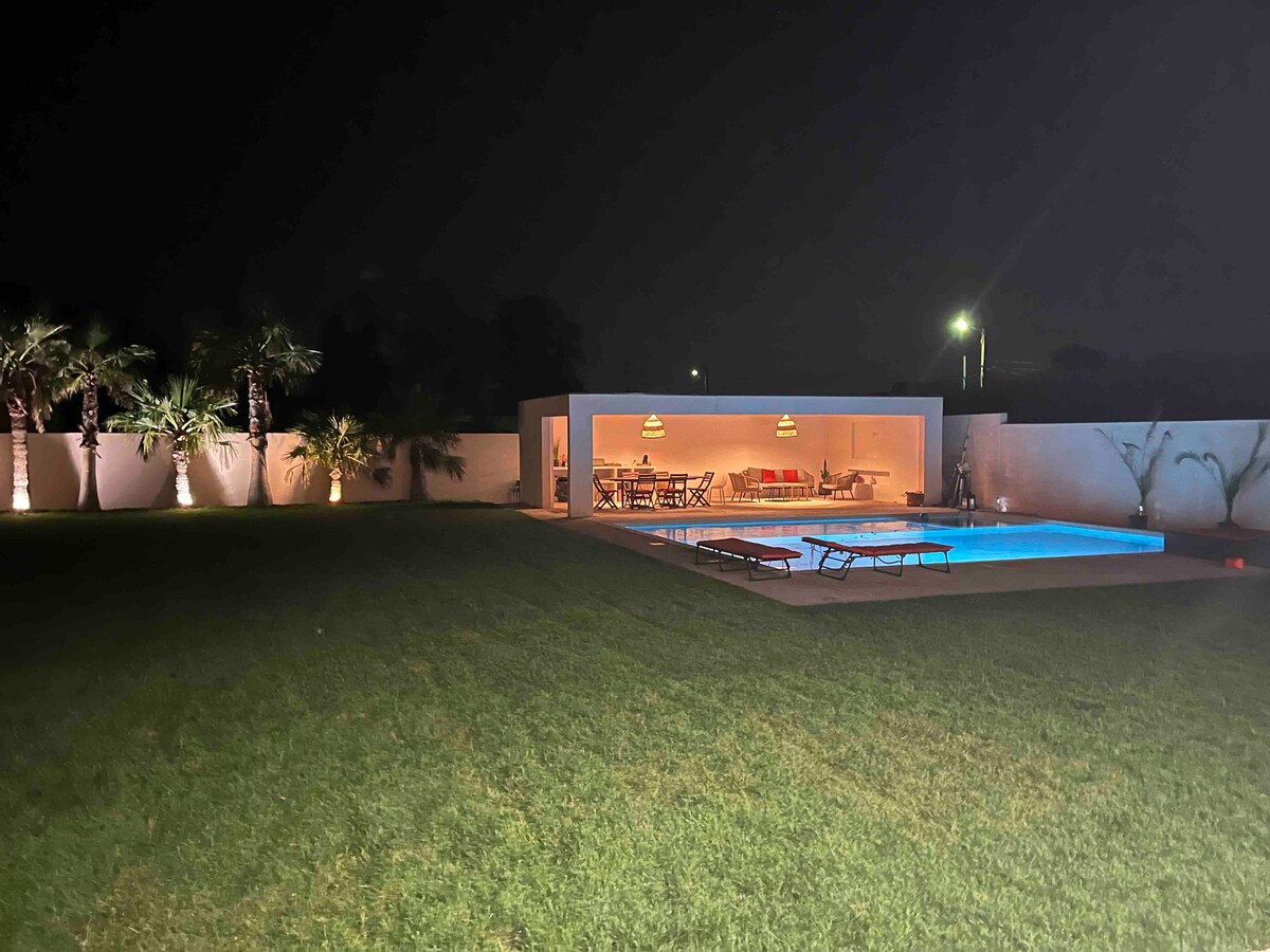 The outdoor area features a well-maintained lawn, a large illuminated swimming pool, and a modern pool house with lounge seating. Landscape lighting creates a soft ambiance, highlighting palm trees and garden borders, while the peaceful night sky adds a serene backdrop.