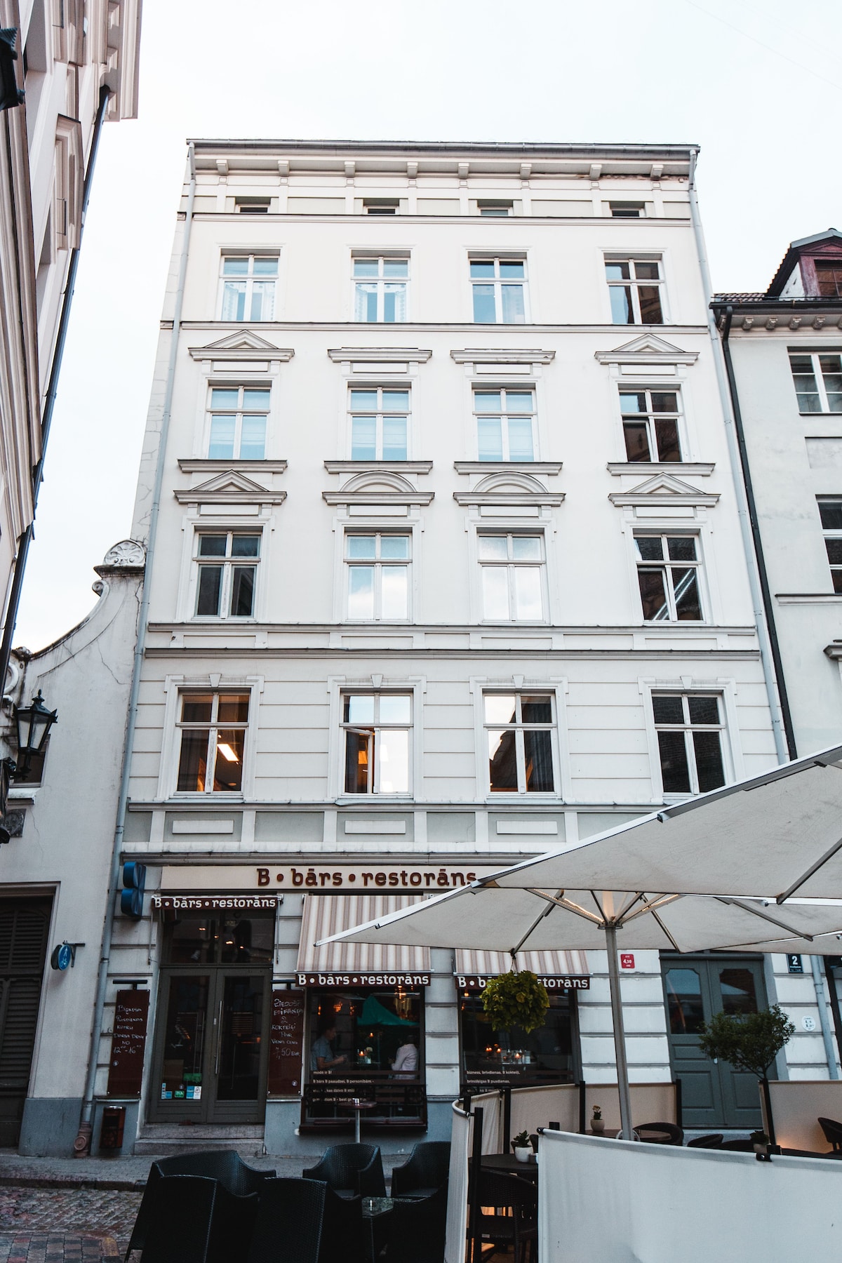 The exterior of a well-maintained four-story building is presented, featuring white walls and multiple symmetrical windows. The ground floor includes a restaurant entrance with tables and chairs on the outdoor patio, contributing to a vibrant street atmosphere.