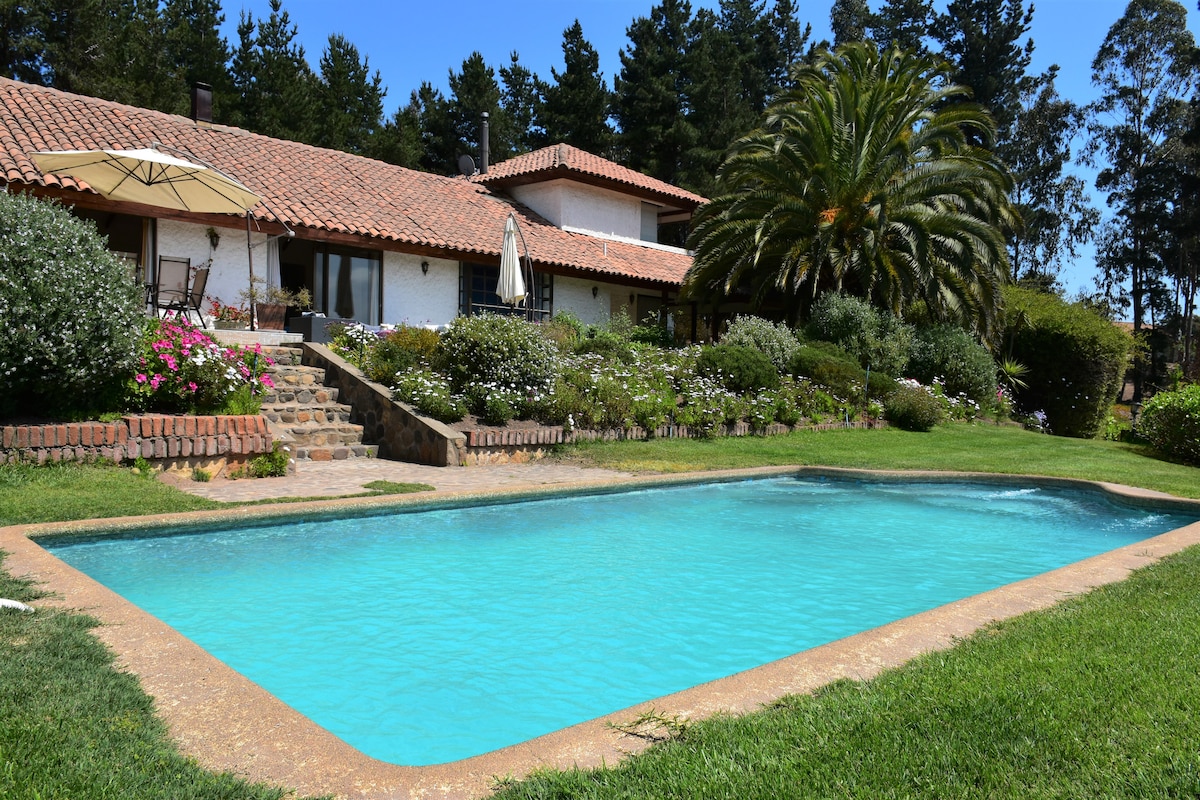 A tranquil swimming pool is set amidst lush greenery and floral gardens. Steps lead from the pool to the charming house, which features a terracotta roof and several outdoor seating areas under umbrellas. Tall trees provide shade around the area.