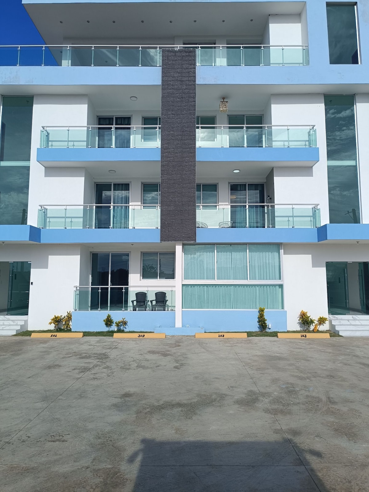 A modern building exterior features multiple balconies with glass railings. The façade showcases blue and white accents, with a vertical dark panel offering contrast. Potted plants are positioned along the ground level, complementing the spacious parking area in front.