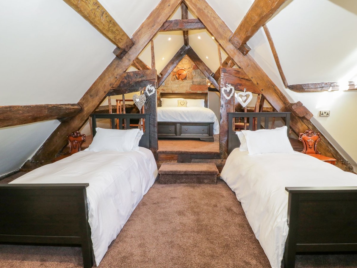 A charming loft bedroom showcases exposed timber beams and subtle lighting. Two beds with white linens are positioned on a plush carpet, with a cozy sleeping area beyond featuring additional furnishings. Warm accents offer a welcoming touch to the rustic space.