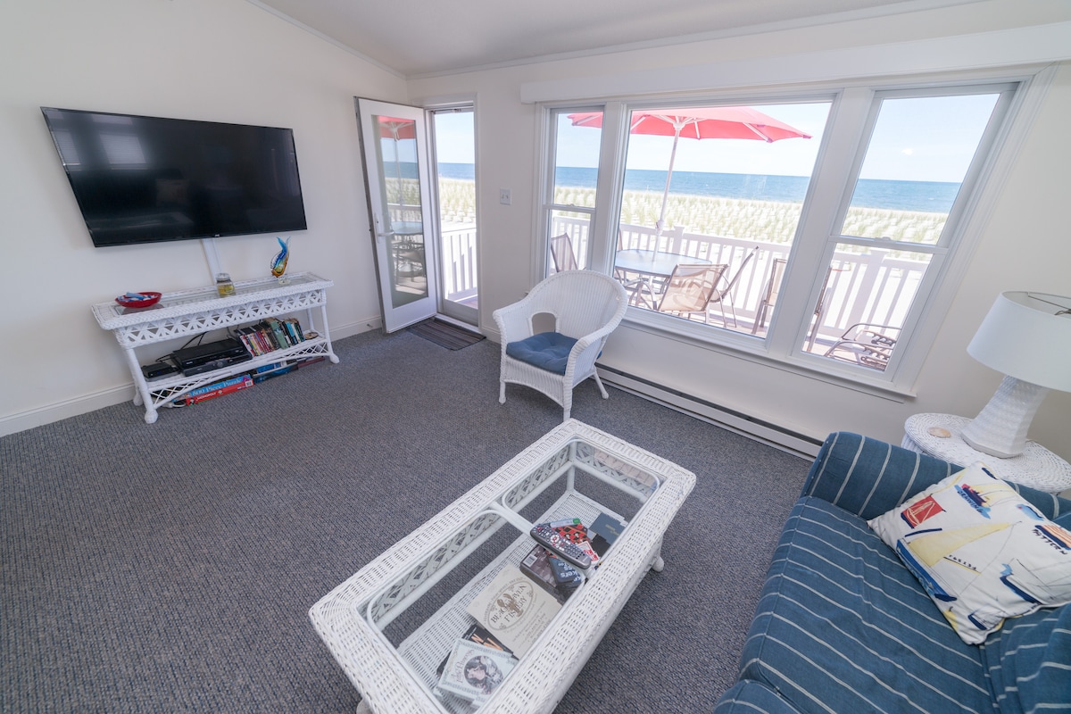 A bright living area features a flat-screen TV mounted on the wall and a cozy blue sofa with decorative pillows. Large windows provide views of the ocean, while a glass coffee table rests on the carpeted floor. A door leads to a private deck with an umbrella.