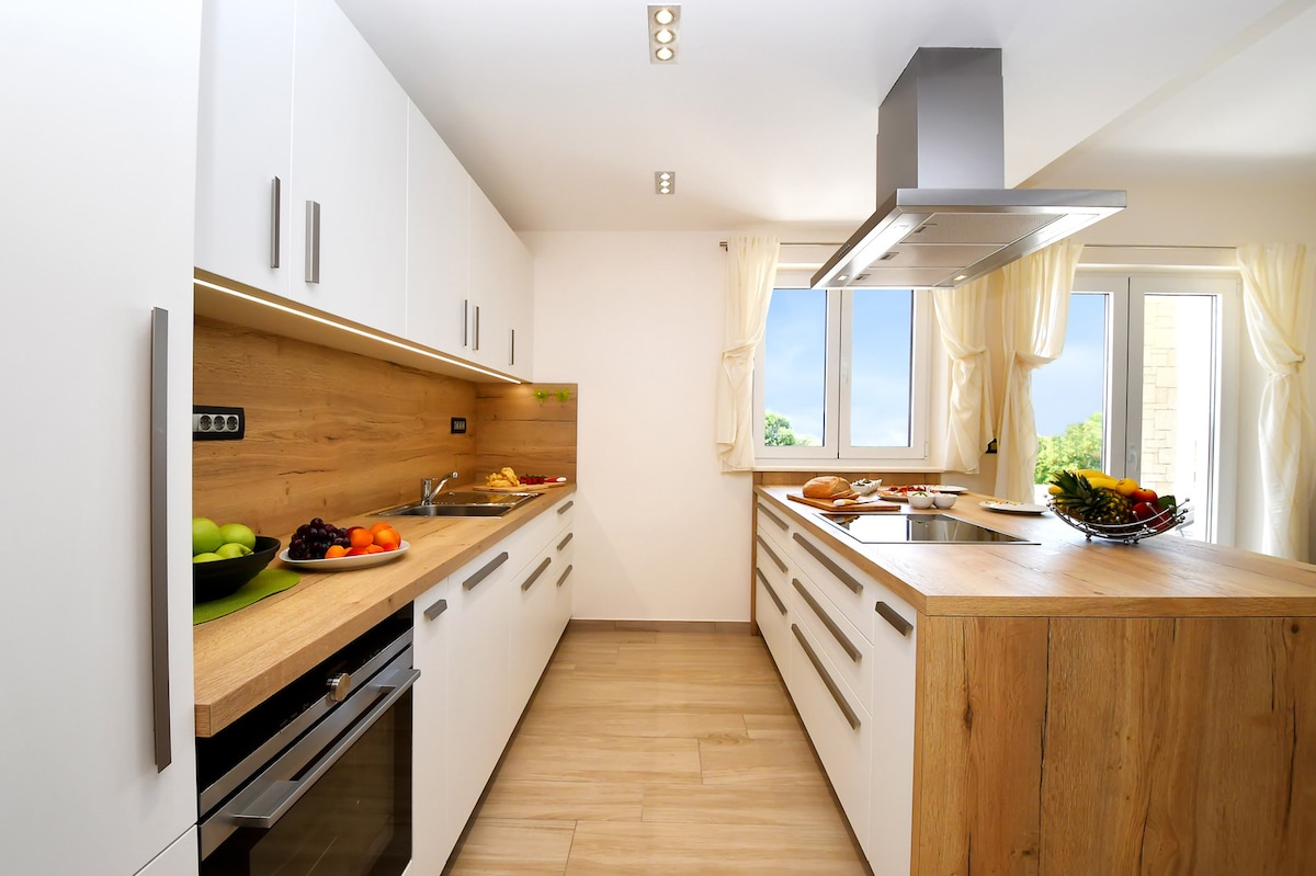 A modern kitchen features sleek white cabinetry paired with natural wood accents. Countertops offer ample space, with various fresh fruits displayed in bowls. Large windows provide natural light, while a stylish range hood is positioned above the cooking area.
