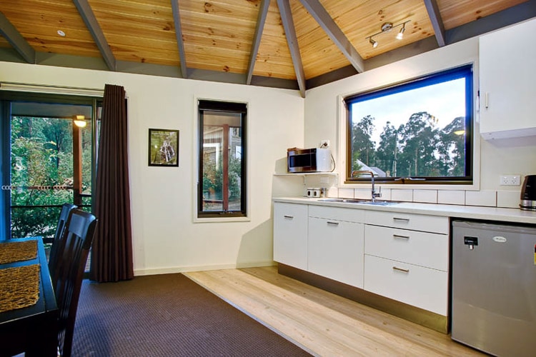 A well-equipped kitchen is visible, featuring sleek white cabinetry, a stainless steel fridge, and a microwave. Large windows provide natural light and views of surrounding greenery. The open layout includes a dining table and wooden flooring, complemented by a high ceiling with exposed beams.