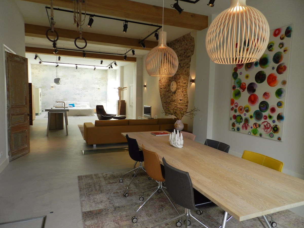 A spacious dining and living area features a long wooden table surrounded by colorful chairs. Two unique pendant lights hang from the ceiling. A textured wall adds visual interest, while a large piece of art with vibrant colors enhances the room's design. Natural light floods the space.