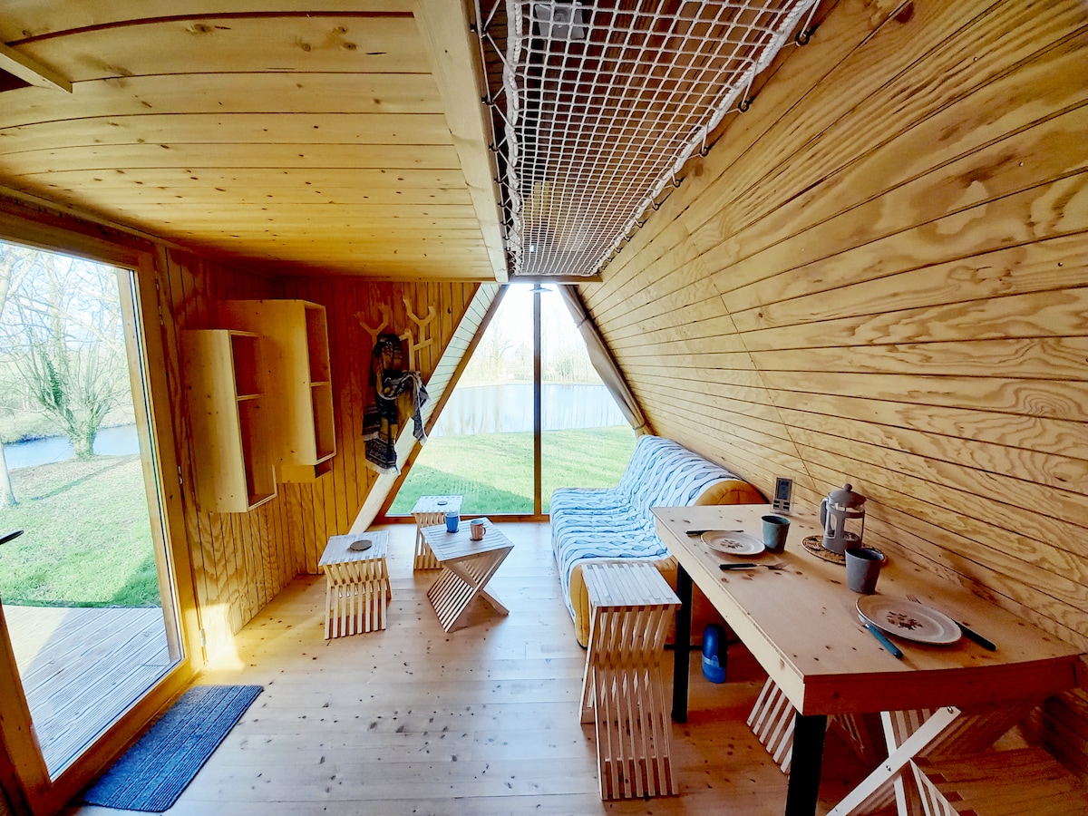 The interior of the cabin features wooden walls and a spacious layout. A cozy seating area is arranged with a sofa and a coffee table. A dining table is visible, accompanied by simple dinnerware. Large windows provide views of the surrounding nature.