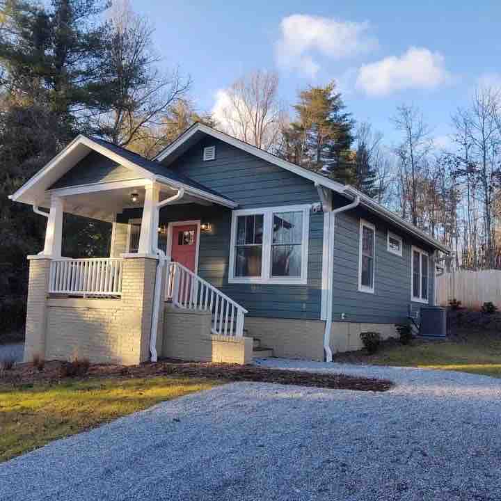 A charming cottage is positioned under a clear blue sky, featuring a light-colored facade with white trim. A covered front porch enhances the entrance, while a gravel path leads to the home. Surrounding greenery adds a natural touch to the setting.