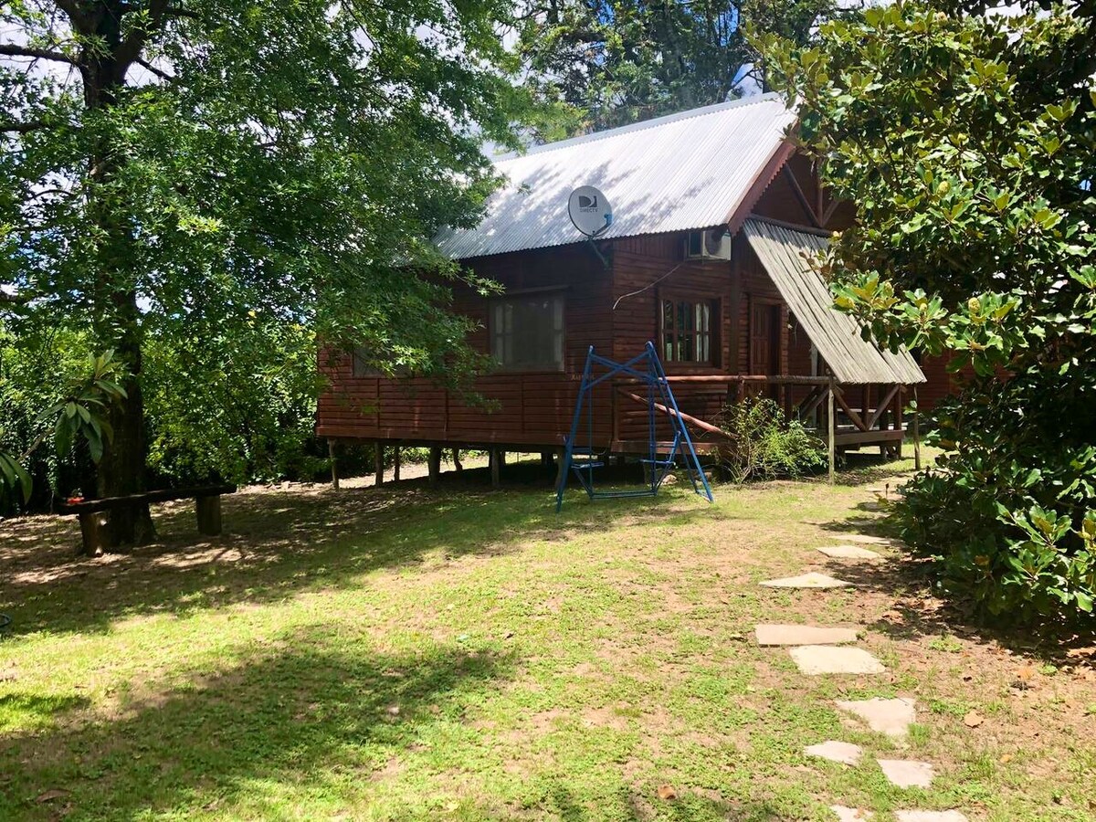 A wooden cabin is positioned slightly elevated on stilts, surrounded by a well-maintained garden. A swing set stands nearby, with stepping stones leading up to the porch. Trees provide ample shade, and an air conditioning unit is mounted on the cabin's exterior.