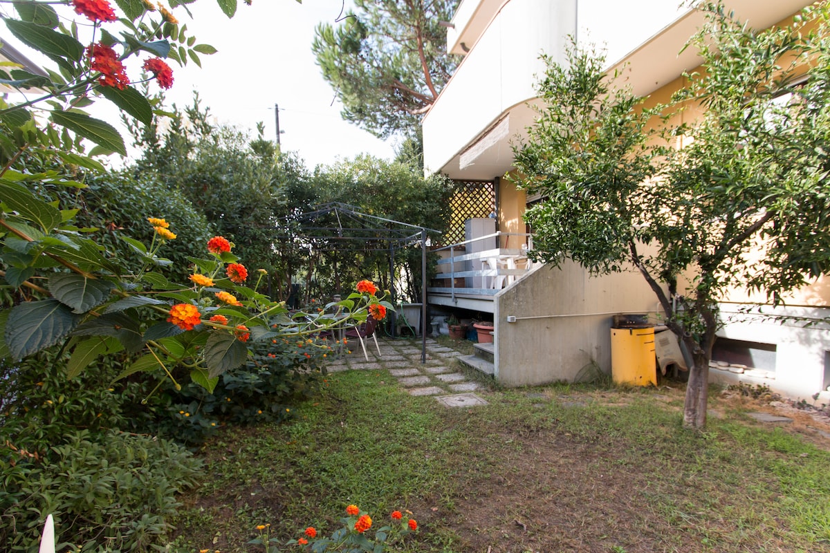 A private garden is surrounded by vibrant flowers and greenery. A stone pathway leads to the entrance of the house, lined with bushes and plants. The area features a small gazebo and a corner with utility storage, providing a tranquil outdoor space.