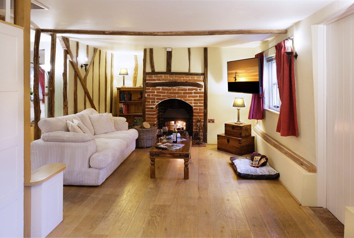 A cozy lounge features exposed wooden beams and a brick fireplace, enhanced by soft lighting from wall sconces. Two comfortable sofas are positioned around a wooden coffee table, with a decorative rug beneath. A TV is mounted above the fireplace, providing entertainment options.