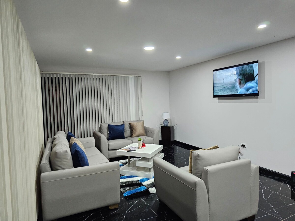 A contemporary living area is furnished with light gray seating arranged around a central coffee table. A wall-mounted television is positioned above the table. Natural light filters through vertical blinds, enhancing the modern aesthetic of the black and white marble floor.