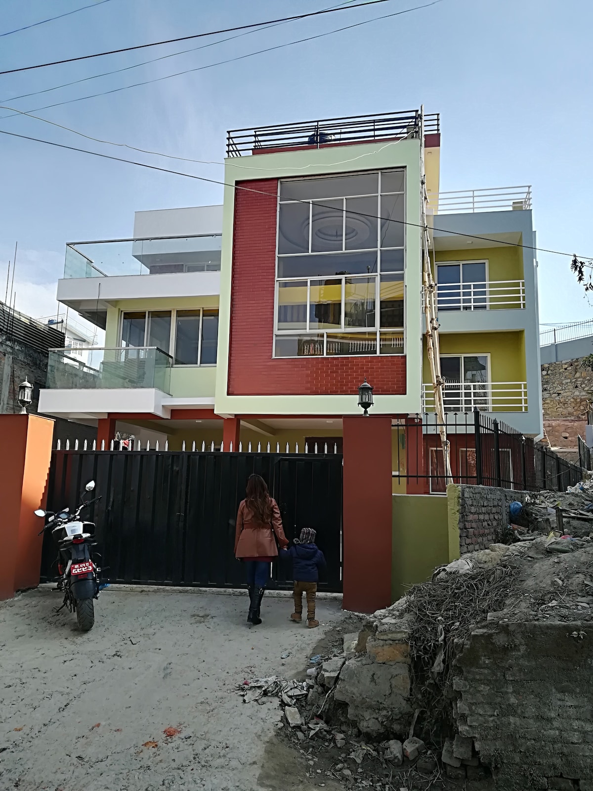 The exterior of a modern bungalow is displayed, featuring a combination of yellow and red accents. A smooth driveway leads to a gated entrance, with a motorcycle parked nearby. A person and a child are approaching the entrance, framed by greenery and a bright sky.